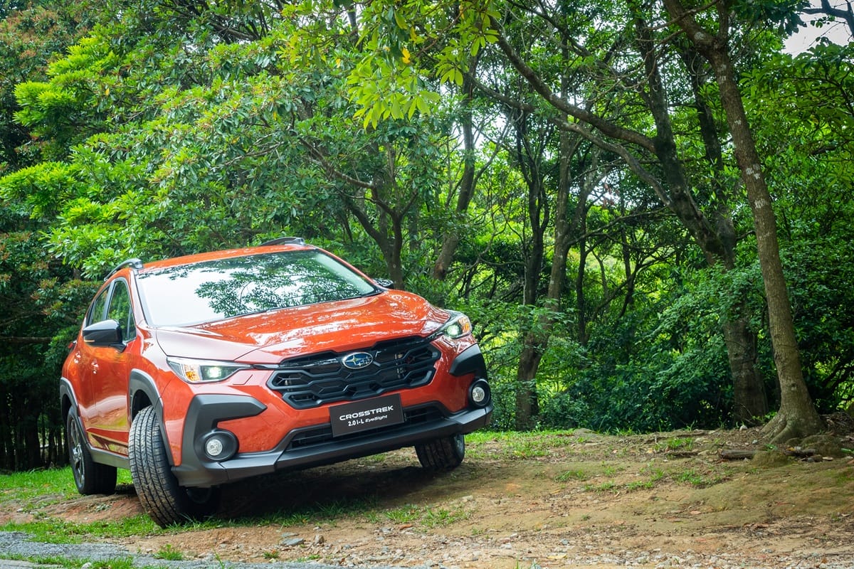 Orange Subaru Crosstrek with LED headlights engaged, climbing a sloped forest trail surrounded by dense green trees and natural terrain.