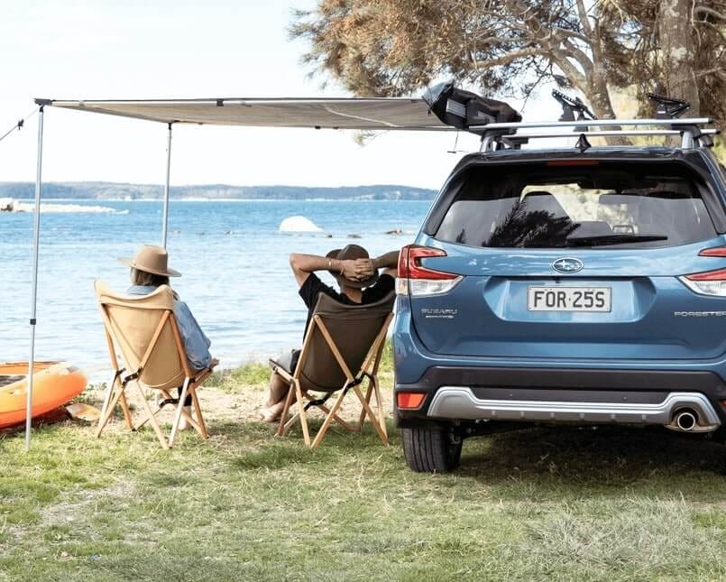 Blue Subaru Forester parked lakeside with two people relaxing under an awning, showcasing a beachside camping setup in Western Australia.