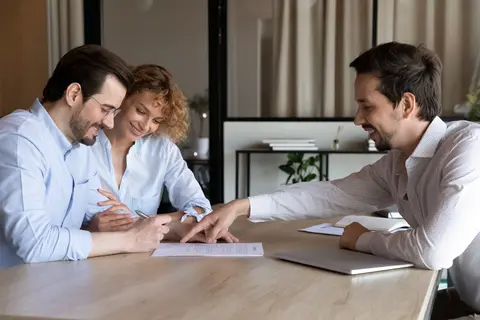 A smiling couple signing a document at a table while a professional points to the paperwork during a meeting