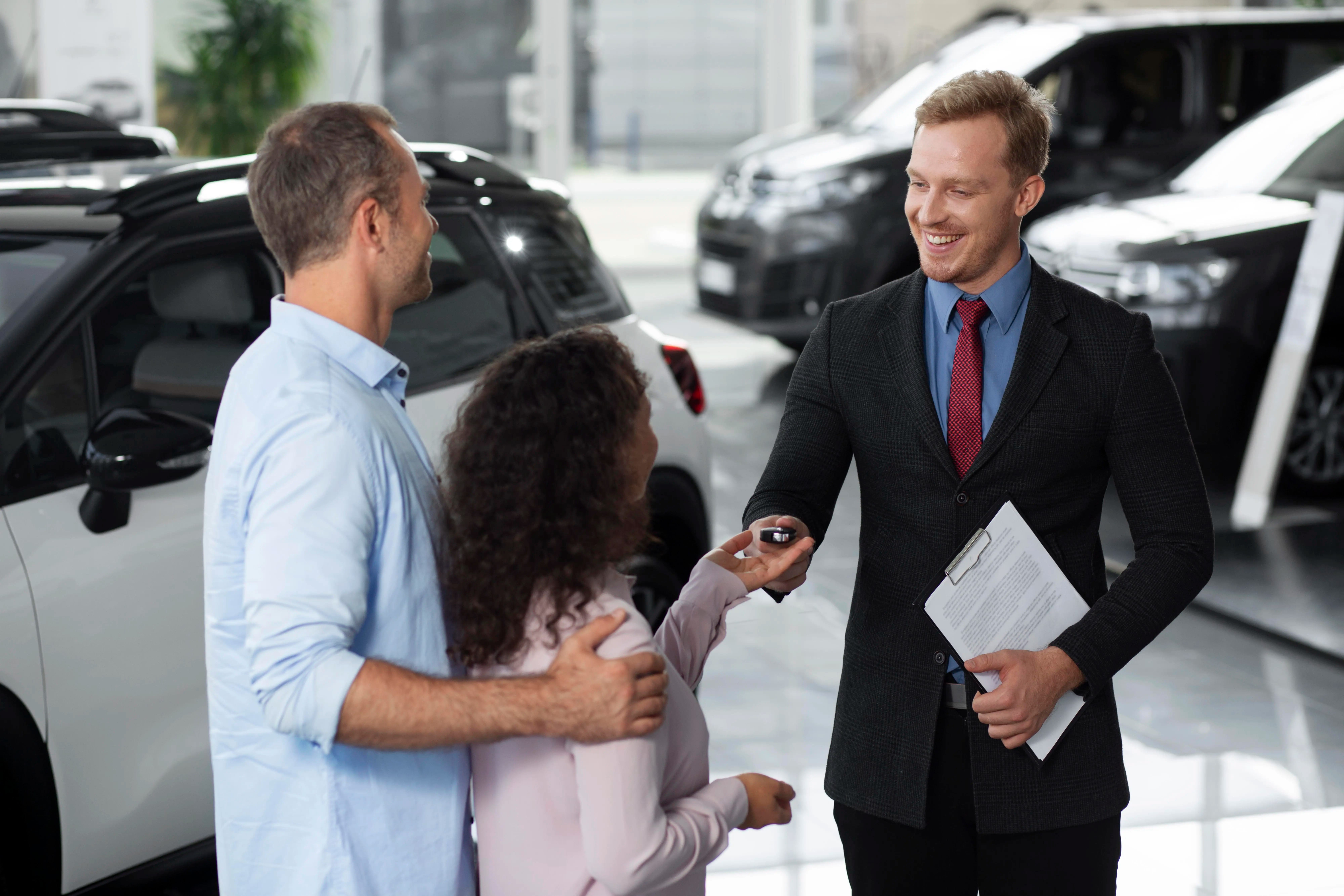 Smiling car dealer handing over keys to a happy couple inside a modern showroom, symbolising a new vehicle purchase experience