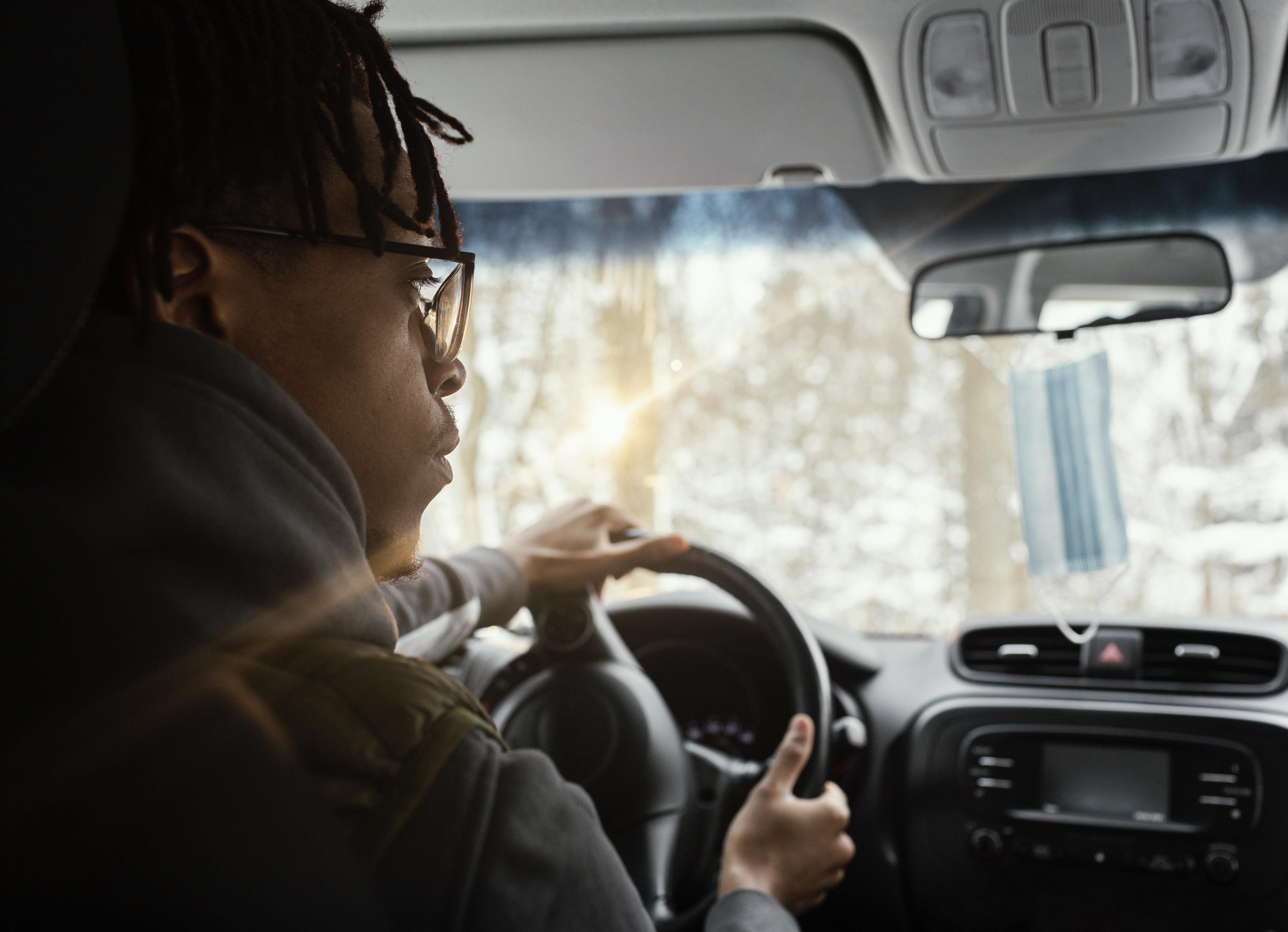 A young man wearing glasses drives a vehicle through a snowy landscape, hands firmly on the steering wheel. 