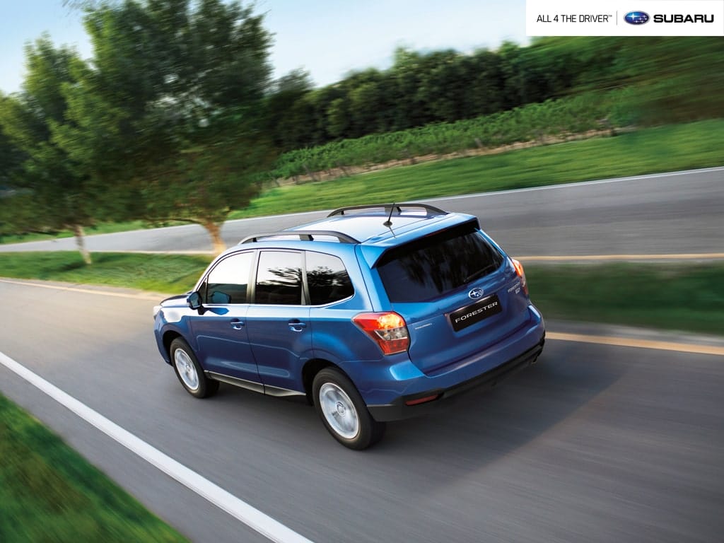A blue Subaru Forester driving along a smooth country road lined with trees and farmland, captured from a rear angle.