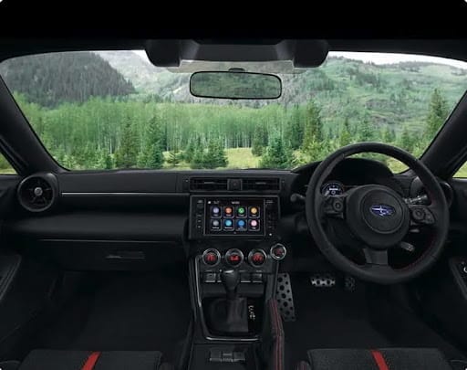 Interior of a Subaru BRZ showing the driver-focused dashboard and infotainment system, with a mountain forest visible through the windscreen