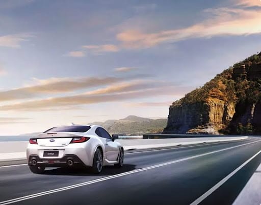 White Subaru BRZ driving along a coastal highway with rocky cliffs and a cloudy sky in the background.