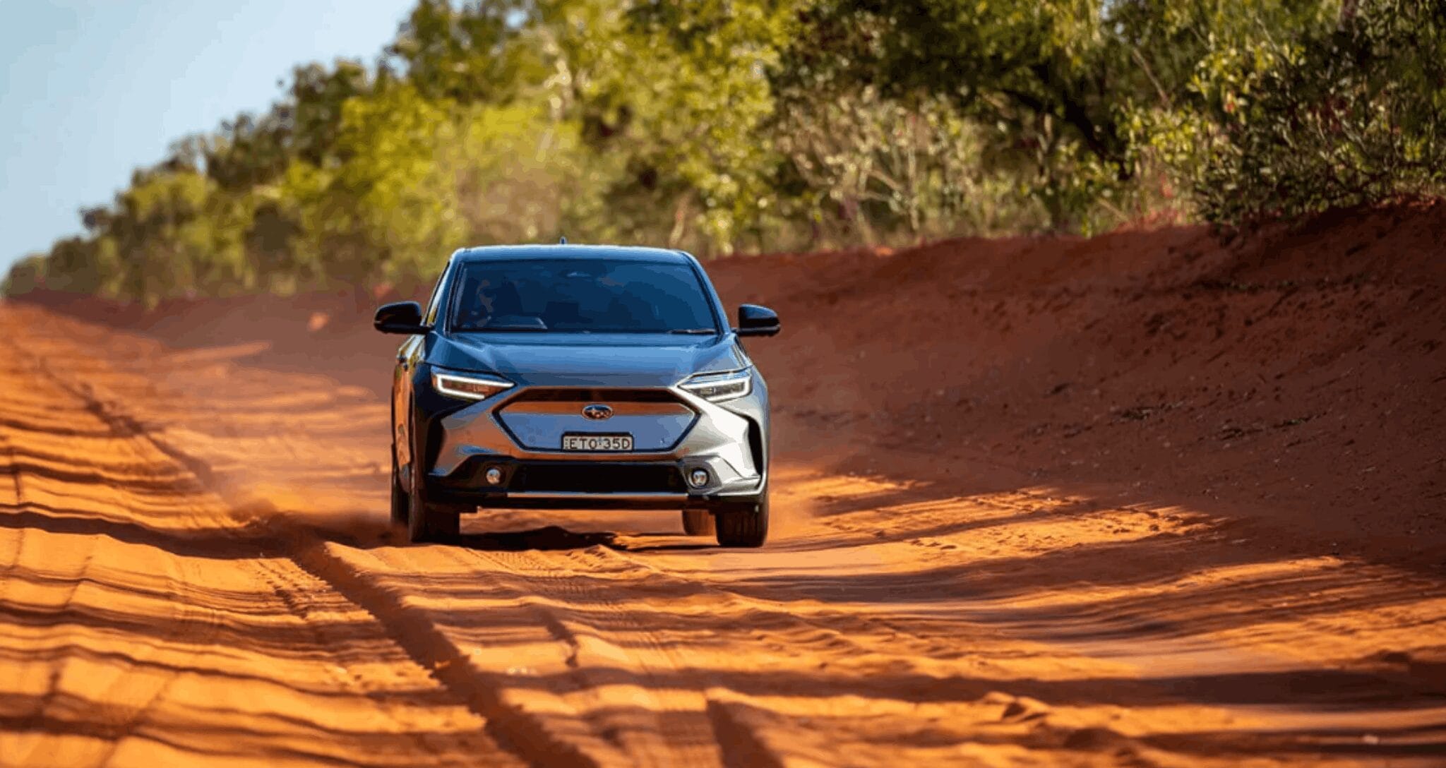 Subaru SUV driving on a red dirt road with dust trails, surrounded by bushland in bright daylight.