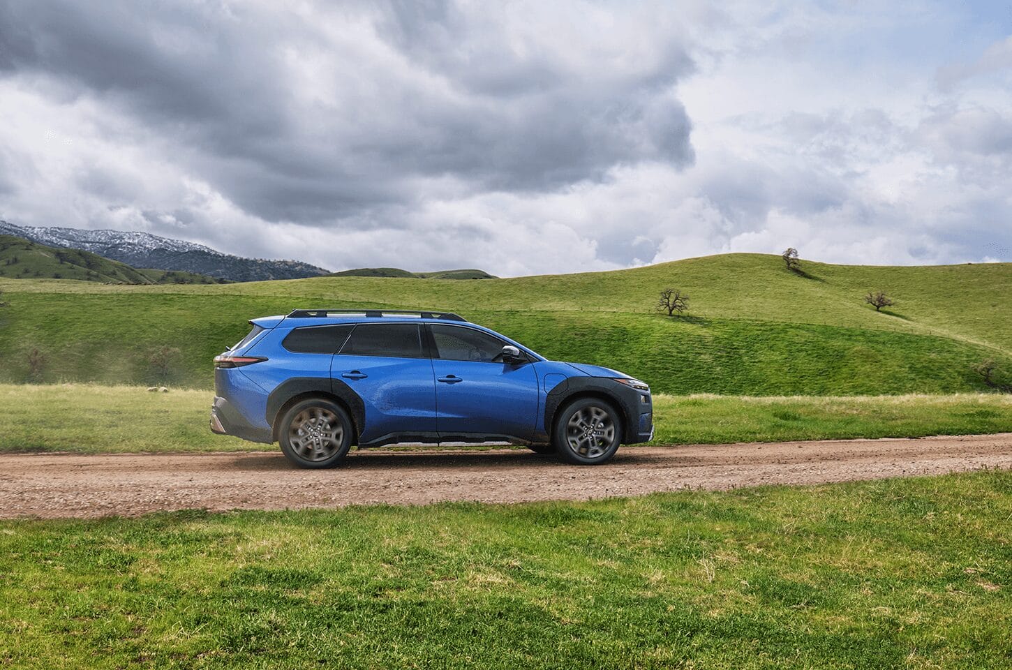 Side profile of the blue Subaru Trailseeker driving through open countryside beneath cloudy skies.]