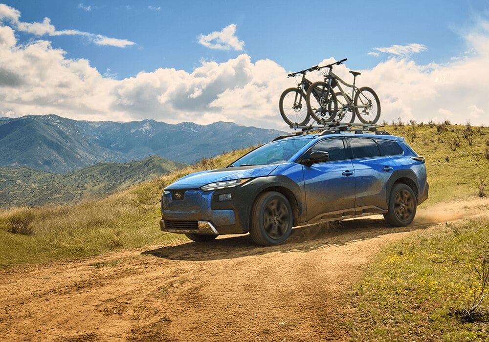 Blue Subaru Trailseeker driving on a dirt track with mountain views and bicycles mounted on the roof.